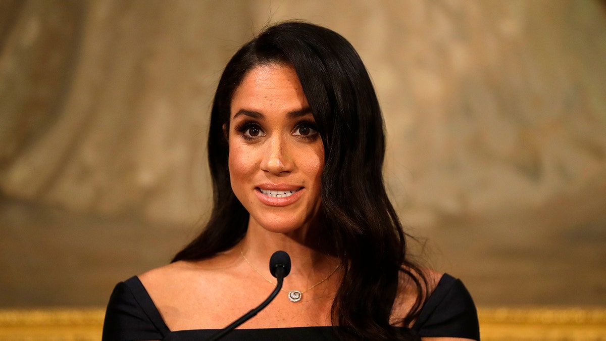 A close-up of Meghan Markle wearing a black dress and speaking to a mic.