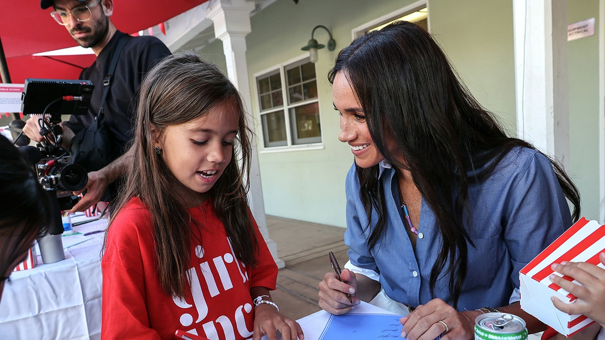 Meghan Markle wearing a denim blouse looking at a young girl wearing a red shirt